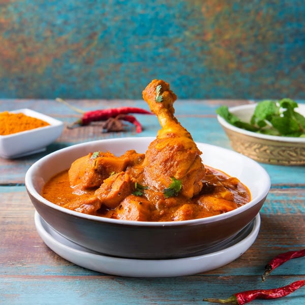 Rasoi's Indian chicken curry with drumsticks in a bowl, spices, and herbs on a blue wooden table.