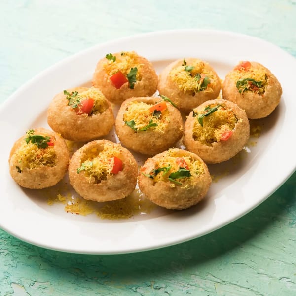 A white platter holds nine golden-brown Indian pani puri, filled with yellow sev, red tomatoes, and green herbs.